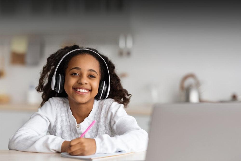 Young girl in a white sweater holding a notebook and pencil and looking at the camera smiling while taking a class at Alabama Connections Academy. 